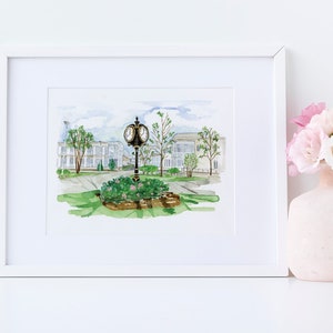 May include: A watercolor painting of a clock tower in front of a row of white buildings. The clock tower is surrounded by green grass and bushes. The painting is framed in a white frame.