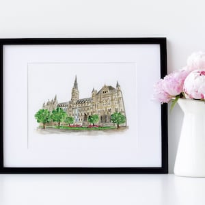 May include: A framed watercolor painting of a large, detailed building with a tall tower. The building is painted in shades of brown and beige, with green trees in front. The painting is framed in black and is displayed on a white surface.