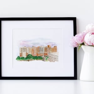 May include: A framed watercolor painting of a city skyline with buildings in shades of brown, red, and green. The painting is set against a light blue sky with white clouds. The frame is black and the mat is white.