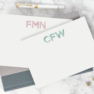 May include: Two white stationery note cards with the initials "FMN" in pink and "CFW" in blue-green. The cards are stacked on top of each other and are on a marble surface.