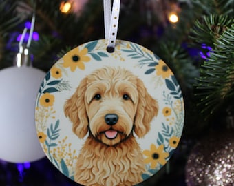 Może przedstawiać: Okrągła ozdoba świąteczna z portretem psa golden doodle. Ozdoba ma białe tło z kwiatową obwódką z żółtych kwiatów i zielonych liści. Wisi na białej wstążce.
