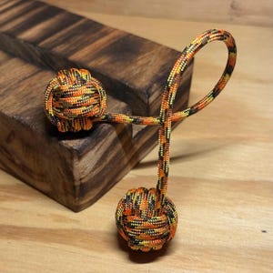 May include: A decorative knot made of orange, yellow, and black paracord. The knot is in the shape of two balls connected by a loop. The item is displayed on a wooden surface.