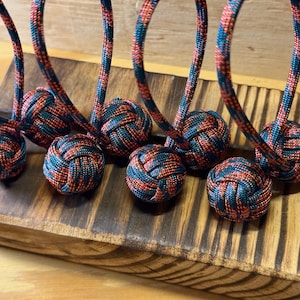 May include: Close-up of several decorative knot balls with matching cords. The balls and cords are a combination of red, navy blue, and teal. The objects are resting on a wooden surface.