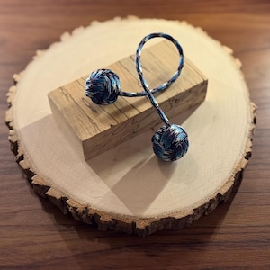 May include: A decorative wooden block with a multi-colored corded object. The corded object has two spherical ends with blue, white, and black patterns. The block is resting on a round, natural wood slice with a rough bark edge.