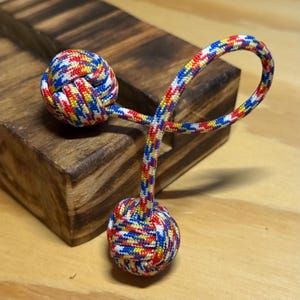 Może przedstawiać: Ozdobny węzeł, z żywą mieszanką czerwonych, żółtych, niebieskich i białych sznurków. Węzeł jest połączony z pętlą i innym węzłem, wszystko w tej samej kolorystyce. Przedmiot jest umieszczony na drewnianym tle.