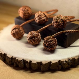 May include: Close-up of several decorative copper-colored ball-shaped objects with long, thin stems. The objects are arranged on a dark wooden block and a light-colored wooden surface. The balls have a textured, woven appearance.