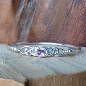 May include: A silver bangle bracelet featuring a central oval-cut light purple amethyst gemstone. The metal band displays intricate knotwork detailing on both sides of the stone, with smooth, rounded silver accents at the connection points.