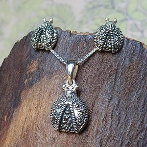 Peut inclure: Un ensemble de bijoux en argent composé d'un collier avec pendentif et de deux boucles d'oreilles assorties. Chaque pièce présente un motif de coccinelle incrusté de petites pierres de marcassite sombres à facettes.