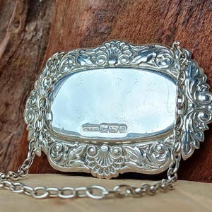 May include: An antique silver bottle label, oval in shape with intricate scrollwork and a chain. The label has a reflective surface and is likely vintage. The hallmarks are visible.