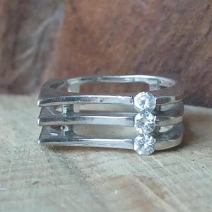 May include: A silver ring with a distinctive design. The ring has three horizontal bands, each set with a clear, round gemstone. The gemstones are evenly spaced, adding sparkle. The ring is set against a neutral background.