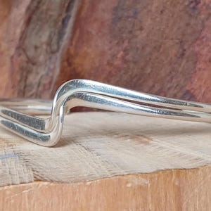 Könnte beinhalten: Ein silberner Ring mit einem einzigartigen Wellendesign. Der Ring hat eine Doppelbandkonstruktion mit glatter, polierter Oberfläche. Der Ring ist auf einer Holzoberfläche platziert, was seinen metallischen Glanz hervorhebt.