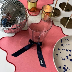May include: A pink table runner with a scalloped edge, a clear glass with a black ribbon tied around it with the name "Jesse" written on the ribbon, a silver disco ball, and a white plate with blue splatter paint.