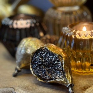 May include: Two golden fig sculptures with black interiors. The figs are sitting on a wooden surface with a candle holder in the background.