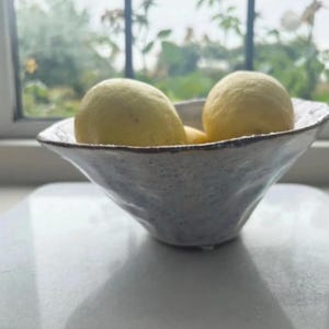 May include: A ceramic bowl with a textured, light blue and white exterior holds several yellow lemons. The bowl has a unique, organic shape and sits on a white surface, with a window and greenery in the background.