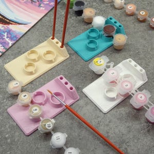 May include: An overhead shot of paint-by-numbers supplies. The image shows several paint palettes in various colors, including pink, teal, white, and beige. Small paint pots are arranged around the palettes, along with a paintbrush and a partially visible canvas.