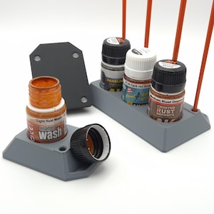 May include: A gray plastic paint bottle holder with several small paint bottles. One bottle is open, displaying a light rust wash enamel. Other bottles include dark red and medium rust deposits. The holder has a unique design.