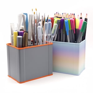 May include: Two rectangular containers filled with art supplies. One is gray with an orange trim, holding brushes, tools, and pens. The other is a pastel ombre, holding colored pencils and markers. The containers are on a white background.