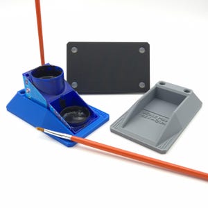 May include: A blue and gray paint station with a black base and a paint brush. The paint station has a blue container with black paint inside. A gray tray is next to the blue container. An orange paint brush is in front of the paint station.
