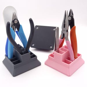 May include: Two sets of jewellery-making tools, each in a different colour organiser. One set includes blue and black pliers, the other set includes pink and orange pliers. A white leather tool holder is in the background.