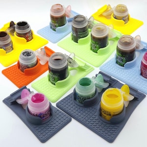 May include: A selection of Citadel paint pots, each resting on a coloured, textured holder. The holders are in yellow, blue, orange, green, and grey. The paint pots contain various colours of paint, ideal for miniature painting.