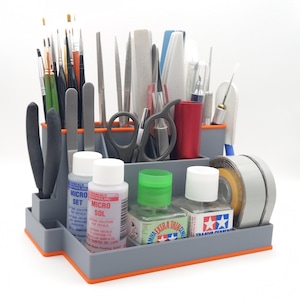 May include: A grey plastic model-making organiser with orange trim, filled with various tools, including brushes, files, scissors, and bottles of paint and solvents. The organiser includes bottles labelled "MICRO SET" and "MICRO SOL".