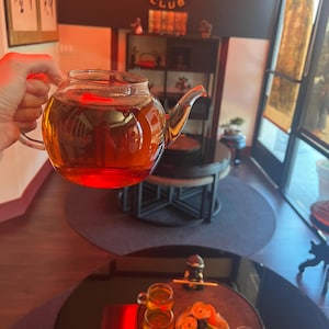 May include: A clear glass teapot filled with amber tea is held up in front of a restaurant interior. The restaurant's sign reads "GOLDEN DRAGON CLUB". Small teacups and a plate of fruit are on a table.