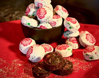 May include: A dark brown bowl filled with multiple round tea cakes wrapped in white paper featuring a red rose illustration and Chinese text. Several additional unwrapped, compressed dark brown tea cakes are placed in front of the bowl.