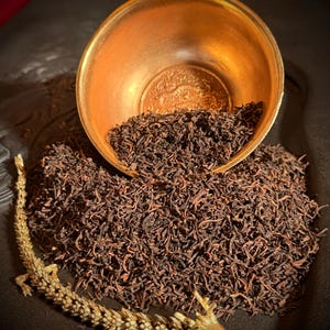 May include: A metallic gold-toned bowl with an embossed dragon design on the interior, partially filled with loose dark brown tea leaves. A pile of the same tea leaves sits in front of the bowl, accompanied by a gold-toned dragon figurine.