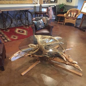 May include: A rustic coffee table made from a large piece of driftwood with a glass top. The table has a natural, organic look and is perfect for a living room or den.