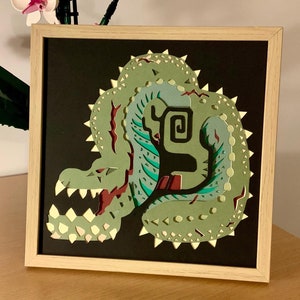 Peut inclure: Une œuvre d'art encadrée en papier découpé représentant un monstre vert et sarcelle sur fond noir. L'œuvre est dans un cadre en bois clair, exposée sur une surface en bois. Le monstre a un aspect détaillé et texturé.
