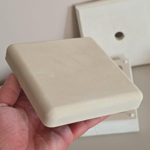Pode incluir: Um molde quadrado de cerâmica em tom branco sujo com bordas arredondadas e uma superfície lisa e mate. O objeto é plano no topo e apresenta uma construção espessa e sólida. Ao fundo, é visível uma segunda peça de molde a condizer.