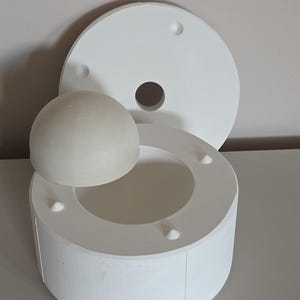 May include: A white ceramic mold set, featuring a round base, a lid with a central hole, and a hemispherical form. The set is designed for crafting or molding, with a clean, minimalist aesthetic. The pieces are arranged on a light-colored surface.
