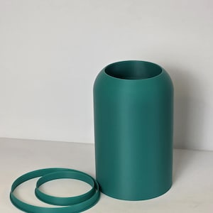 Puede incluir: Un jarrón cilíndrico de color verde azulado con una parte superior redondeada, acompañado de dos anillos verde azulado a juego. El jarrón es de color liso y se mantiene erguido. Los anillos se colocan cerca del jarrón.