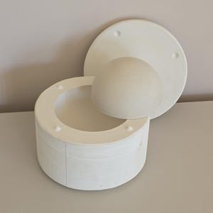 Puede incluir: Un juego de moldes de cerámica blanca, compuesto por una base cilíndrica, una forma hemisférica y una tapa circular. La tapa y la base tienen pequeños agujeros. Los objetos están dispuestos sobre una superficie gris claro.