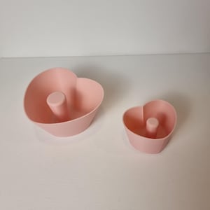 May include: Two pink heart-shaped silicone cake molds. The larger mold is in the background and the smaller mold is in the foreground.