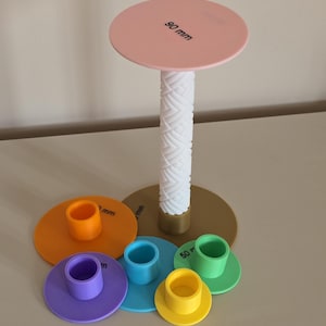 Könnte beinhalten: Ein vertikaler Ständer mit einer weißen, strukturierten Säule und einer rosa runden Platte mit der Aufschrift 90 mm. Um die Basis herum befinden sich fünf kleinere runde Scheiben in Orange, Lila, Blau, Gelb und Grün mit Öffnungen.
