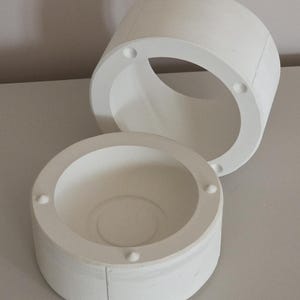 May include: Two-part white cylindrical object with a circular opening. The top piece is a ring with four evenly spaced holes. The bottom piece is a bowl shape with a central indentation and four small, evenly spaced protrusions.