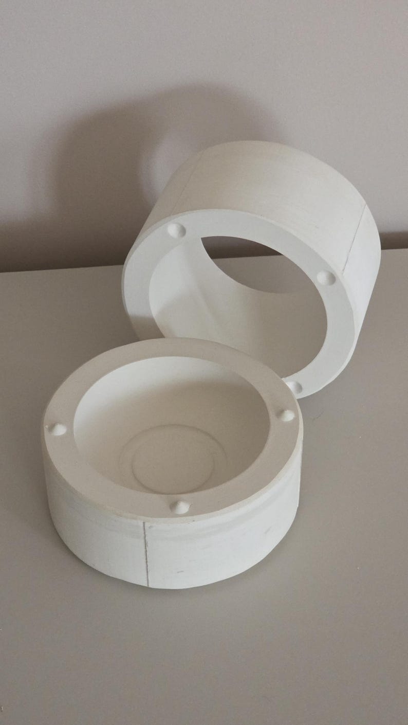 May include: Two-part white cylindrical object with a circular opening. The top piece is a ring with four evenly spaced holes. The bottom piece is a bowl shape with a central indentation and four small, evenly spaced protrusions.
