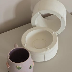 May include: A lilac ceramic mug with floral decorations and a white ceramic mould. The mug has a dark interior and a handle. The mould is in three parts, with a circular opening.