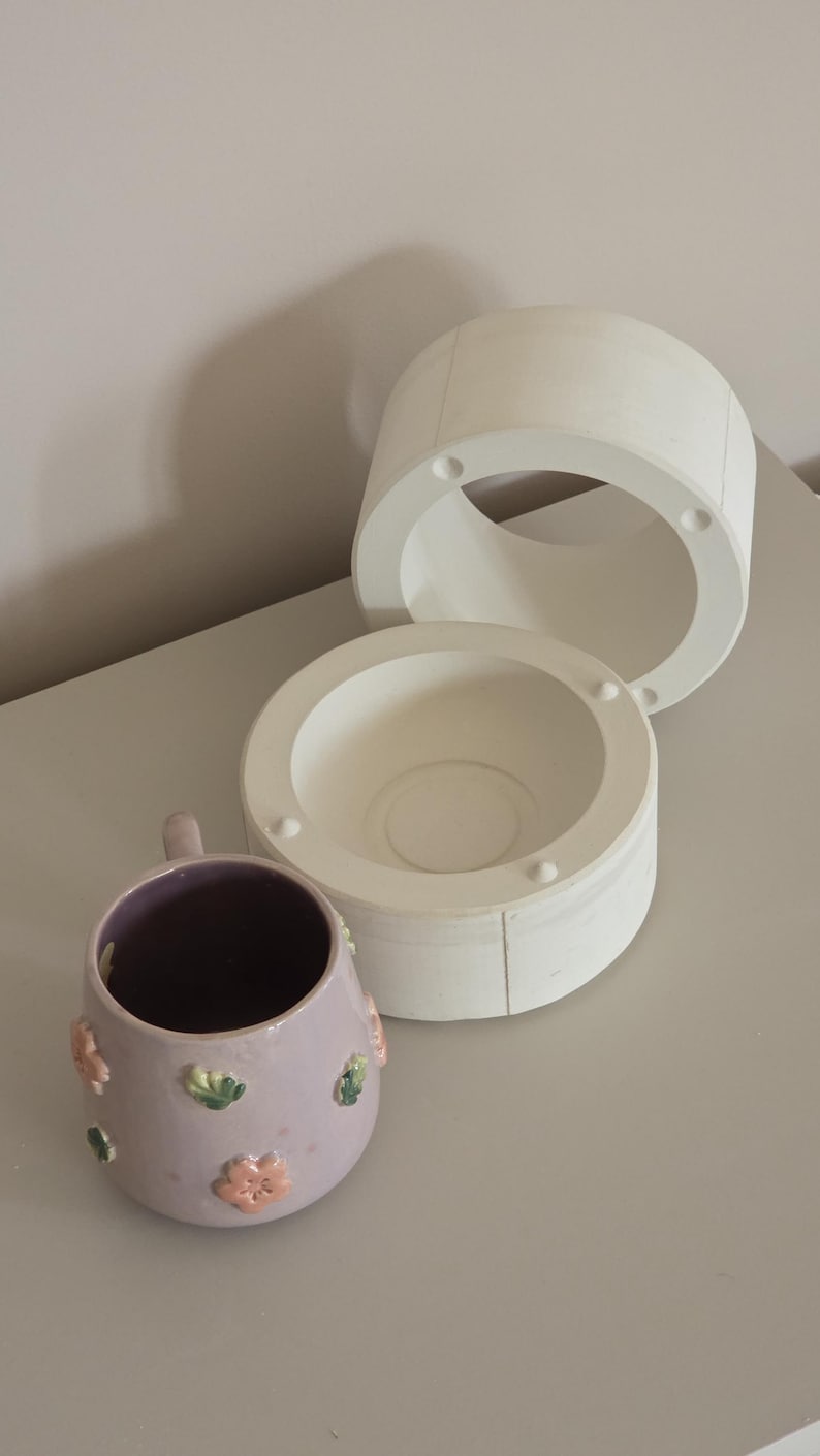 May include: A lilac ceramic mug with floral decorations and a white ceramic mould. The mug has a dark interior and a handle. The mould is in three parts, with a circular opening.