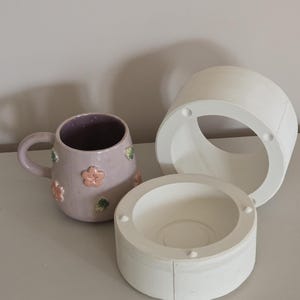 May include: A lavender ceramic mug with pink flower and green leaf decorations, alongside two white ceramic moulds. The mug has a handle and a purple interior. The moulds are cylindrical.