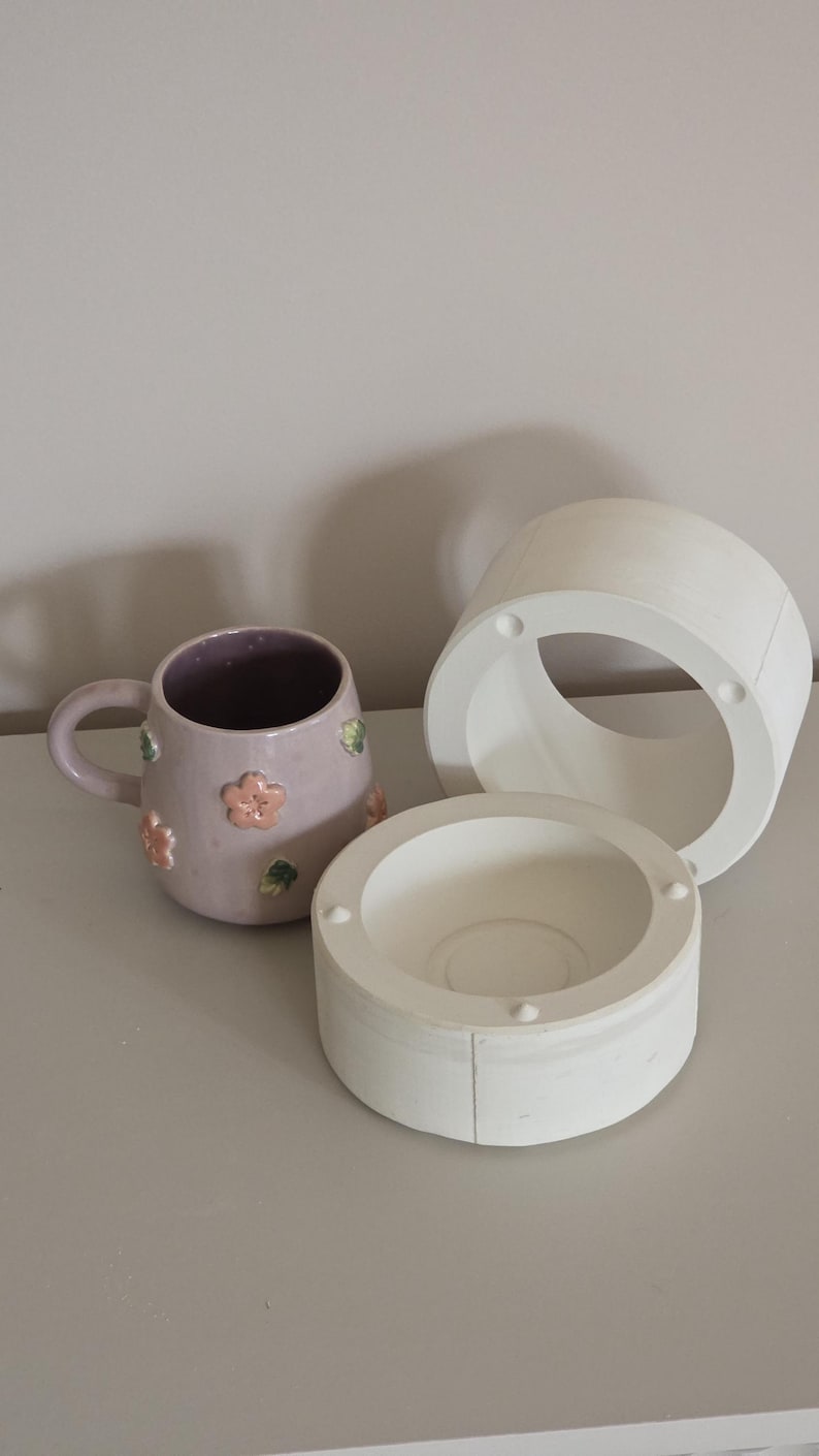 May include: A lavender ceramic mug with pink flower and green leaf decorations, alongside two white ceramic moulds. The mug has a handle and a purple interior. The moulds are cylindrical.