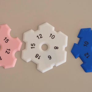 Op de afbeelding: Drie plastic, sneeuwvlokvormige breinaalddiktemeters in roze, wit en blauw. Elk heeft een centraal gat en genummerde markeringen van 6 tot 18. Deze gereedschappen worden gebruikt om de maat van breinaalden te meten.