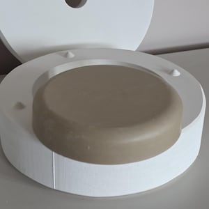 Może przedstawiać: Dwuczęściowa ceramiczna forma, z okrągłą podstawą i pasującą pokrywą. Podstawa zawiera zaokrąglony, brązowy przedmiot. Forma jest biała, z małymi wypukłymi detalami i centralnym otworem w pokrywie. Przedmioty znajdują się na neutralnej powierzchni.