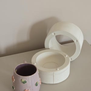 Könnte beinhalten: Eine lila Keramiktasse mit rosa und grünen Blumenverzierungen. Daneben befinden sich zwei weiße Keramikformen, eine zylindrische und eine schalenförmige, wahrscheinlich für Keramikarbeiten. Die Gegenstände stehen auf einer hellgrauen Oberfläche.