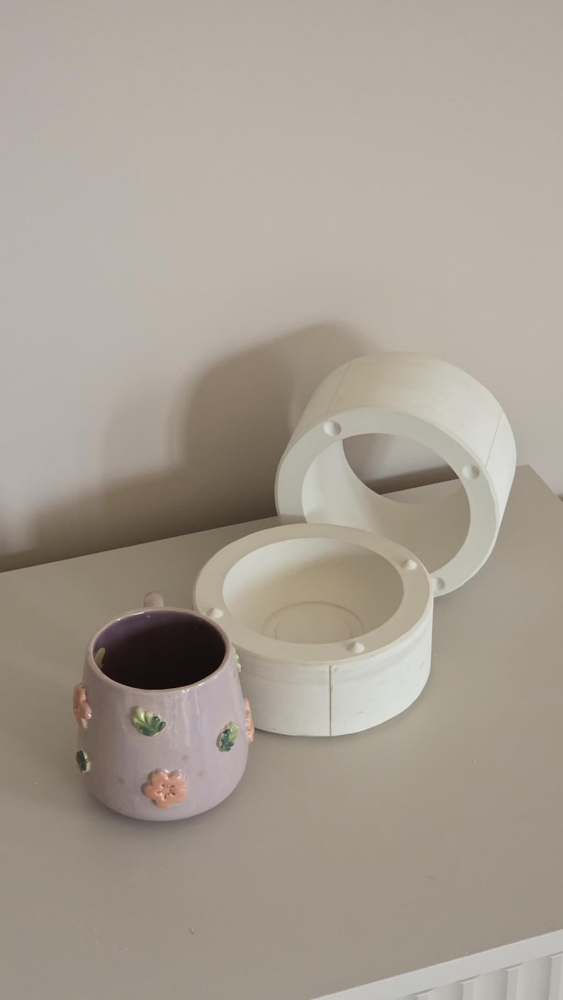 May include: A lilac ceramic mug decorated with pink and green floral designs. Next to it are two white ceramic moulds, one cylindrical and one bowl-shaped, possibly for pottery. The items are on a light grey surface.