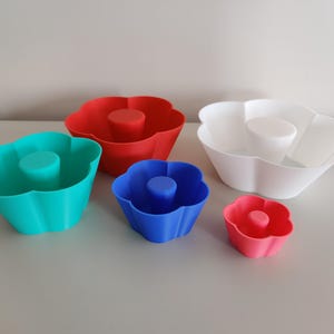 Peut inclure: Ensemble de cinq moules à pâtisserie en silicone en forme de fleur. Les moules sont de différentes tailles et couleurs : rouge, blanc, bleu et turquoise.