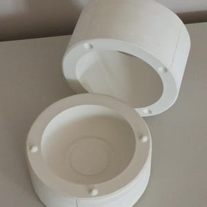 May include: Two-part white cylindrical container set. The top piece is open, revealing the interior. The bottom piece is a shallow bowl shape. Both pieces have small, evenly spaced circular indentations around the rim.