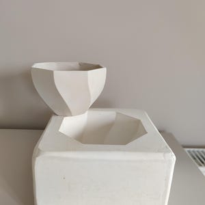 Peut inclure: Pot en céramique blanche avec un design géométrique. Le pot a une base carrée et un dessus hexagonal. Le dessus du pot est conçu pour contenir un bol hexagonal plus petit.