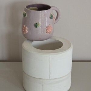 May include: A lavender ceramic mug with a handle, decorated with raised pink and green floral designs. The mug is placed on a white, cylindrical, two-tiered stand. The background is a neutral grey.